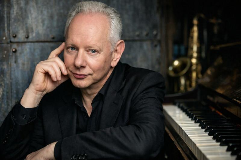 Joe Jackson am Klavier sitzend, Porträt im 16:9-Format mit dunklem Hintergrund und Saxofon im Hintergrund