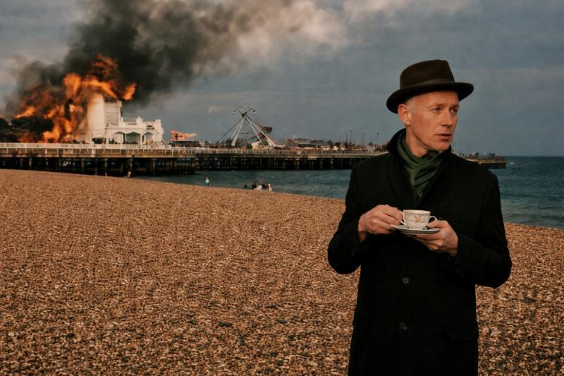 Plattencover von Hope and Fury: Joe Jackson mit Hut trinkt Tee am Kiesstrand, während im Hintergrund ein Piergebäude in Flammen steht
