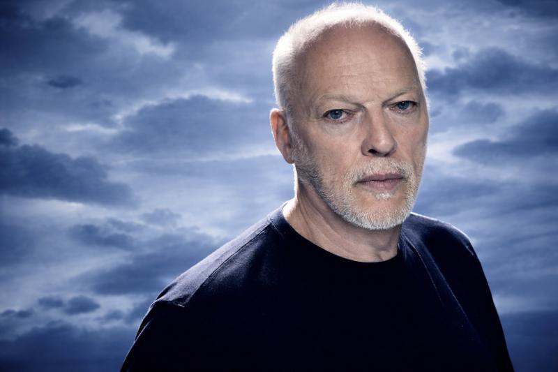 Porträt von David Gilmour mit kurzem weißem Haar und Bart, der ernst in die Kamera blickt; im Hintergrund ein aufgehellter, wolkiger Himmel in Blautönen.