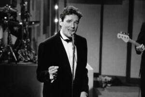 Schwarzweißaufnahme eines eleganten Sängers im Smoking, der vor einem Mikrofon steht und mit ausdrucksstarker Mimik singt; im Hintergrund sind unscharf Instrumente und Bühnenelemente zu erkennen.