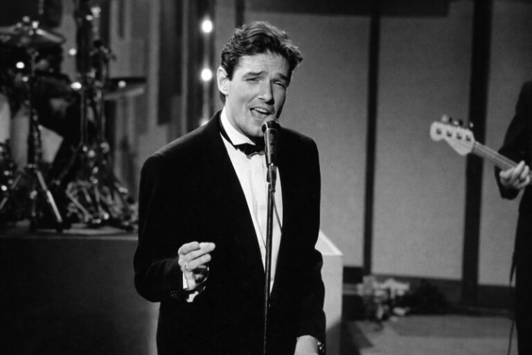 Schwarzweißaufnahme eines eleganten Sängers im Smoking, der vor einem Mikrofon steht und mit ausdrucksstarker Mimik singt; im Hintergrund sind unscharf Instrumente und Bühnenelemente zu erkennen.