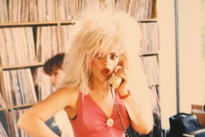 Nina Hagen mit auffälligem blondem Haar und pinkem Outfit telefoniert in einem Raum voller Schallplatten, während im Hintergrund eine weitere Person sitzt.