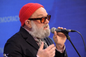 Nahaufnahme von Michael Stipe mit roter Mütze und Sonnenbrille, der in ein Mikrofon singt, vor blauem Hintergrund.