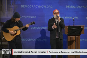Michael Stipe singt ins Mikrofon, während Aaron Dessner links daneben Gitarre spielt, vor einer blauen „Democracy Now!“-Bühnenwand.